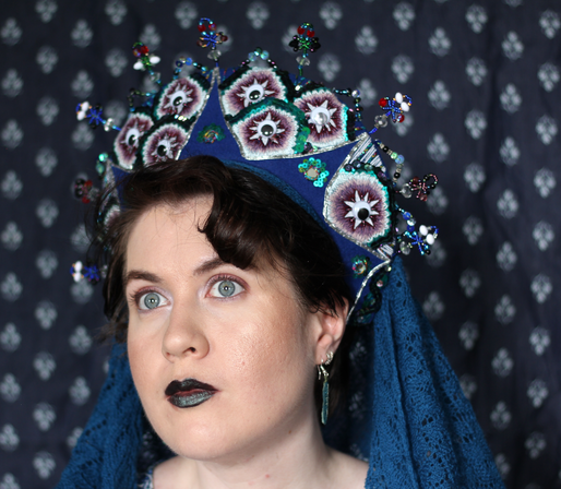 A white woman wearing a blue headdress and a blue veil against a blue patterned background.