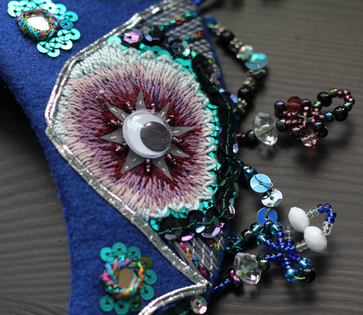 Detail closeup of the embroidery. The middle of the peacock feather is a googly eye surrounded by reflective tape, beading, and embroidery in purple, cream and blue, with sequins at the edging. Above the rim of the headdress there are little beaded wands that’s stand straight up.