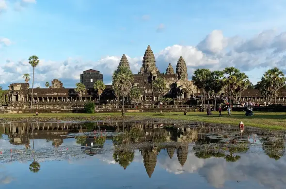 The image features the iconic Angkor Wat temple complex in Cambodia. It is a large religious monument known for its stunning architecture and intricate carvings. The scene depicts the temple surrounded by lush greenery, with a reflecting pool in the foreground that captures the temple's image in the water. The sky shows a mix of clouds, suggesting a beautiful day. The area appears to have visitors exploring the site.

Image Credits: Wikimedia / Jakub Hałun / CC BY-SA 4.0