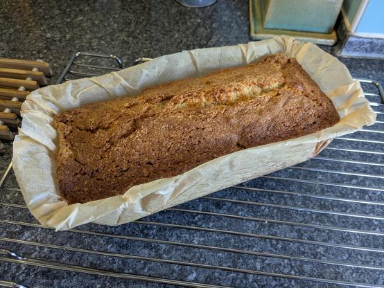 A cake with a browned, cracked top made in a loaf tin with roughly gathered greaseproof paper around the outside, sitting on a shiny steel grille on a dark coloured kitchen work surface