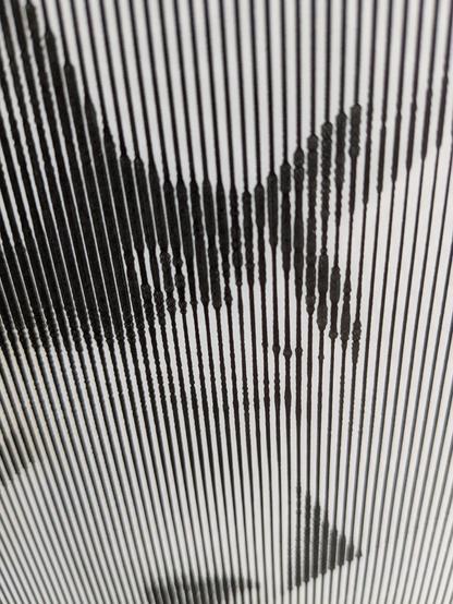 A close up view of a pen plotter drawing that aims to represent a photo of a cat. She's black and white, looking off to the right. The photo is made of lines that are vertical (they look splayed out due to perspective here) and as the photo gets darker the lines spread out, making a wider dark strip