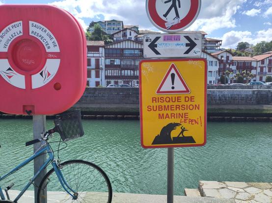 Devant les eaux turquoise du canal portuaire de Saint-Jean-de-Luz, par un bel après-midi ensoleillé, un vélo est accroché à un support de bouée rouge vif. A ses côtés, un panneau sur fond jaune figure une vague gigantesque sur le point d'engloutir une minuscule silhouette et annonce en lettres rouges : "Risque de submersion marine". "Le Pen !" a été ajouté à la main.