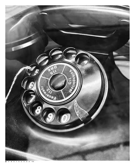 Schwarzweißfotografie: schwarzes glänzendes altes Telefon mit großer Wählscheibe und Hörer, auf der Wählscheibe stehen die wichtigsten Telefonnummern der DDR: Deutsches Rotes Kreuz (DRK) 115, Feuerwehr 112 und Volkspolizei 110