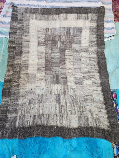 A hand knit afghan-sized blanket is spread out to dry on some beach towels. The blanket is a rectangle made in a spiral, starting with a strip in the middle, which turns each corner at right angles. The colors shift organically from creamy white to dark charcoal gray. Running down the center of each strip is a line of small lace motifs resembling a cat track.
