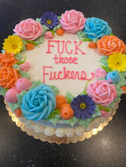 A beautiful round birthday cake with white frosting and big pretty blue and pink and yellow flowers around the edge and in the middle it says in pink serif and cursive letters:

FUCK
those
Fuckers