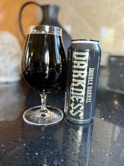 A tall snifter glass of dark beer next to a pint can which is black with silver lettering spelling out “Darkness” on all capitals.
