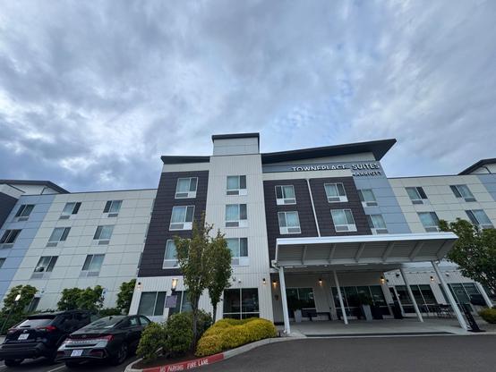 This photo shows the exterior of a modern four-story hotel building under a cloudy sky. The hotel is the TownePlace Suites by Marriott, as indicated by signage at the top. The facade is a mix of light grey, white, and dark grey panels, with many rectangular windows. A white canopy covers the hotel entrance, supported by white columns. The entrance is framed by neatly trimmed shrubs, small trees, and modern outdoor lamps. A fire lane is marked in red at the curb. Several parked cars are visible, including a black SUV and a silver sedan. The perspective is slightly upward, making the building appear tall and structured against the cloudy backdrop.