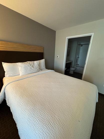 This image shows a neatly made hotel bed in a simple, modern room. The bed features a wooden headboard with a darker strip at the center, white pillows, and a textured white comforter. The lighting is warm, creating a cozy feel. To the right of the bed, a doorway opens into a small bathroom, where a toilet and part of a shower stall with a sliding glass door are visible. The walls are painted in two tones: one side light beige and the other a soft gray. The bed takes up most of the foreground, showing a clean and restful sleeping space.