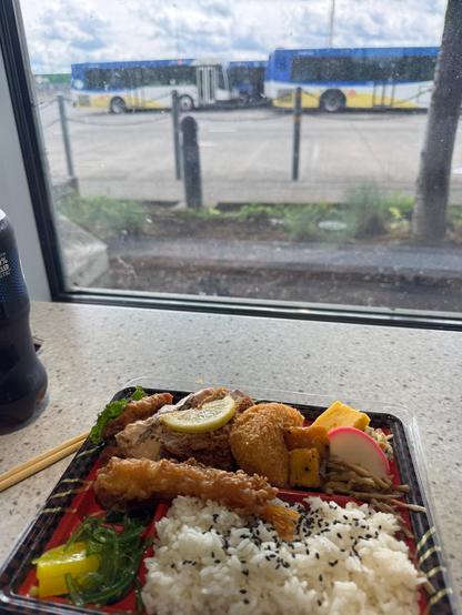 In the foreground, a Japanese-style bento meal sits on a speckled white countertop near a window. The tray contains white rice topped with black sesame seeds, fried shrimp, seaweed salad, yellow pickled vegetables, tamagoyaki (rolled egg), slices of radish, fried croquettes, grilled chicken with a lemon slice, and cooked root vegetables. A pair of wooden chopsticks lies beside the tray, and a soda bottle is partially visible to the left. Through the slightly dirty window, several TriMet buses can be seen parked outside under a cloudy sky, adding a sense of urban and transit-focused atmosphere to the meal scene.