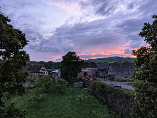 A wide shot captures a collection of rural buildings nestled amongst trees under a dramatic, dusky sky. Several timber-framed buildings with dark roofs are visible, clustered together. A stone wall, partly covered in greenery, runs across the midground, with more trees and a glimpse of a parked car beyond. The sky is a blend of purples, greys, and a streak of orange on the horizon, suggesting either dawn or dusk. The overall scene evokes a sense of quiet countryside at the transition of day and night.
