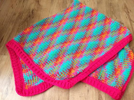 A crochet blanket with an Argyle pattern. The blanket is predominantly a teal colour with the cross cross squares of the pattern in pink and orange. It is sitting on a wooden surface folded up.