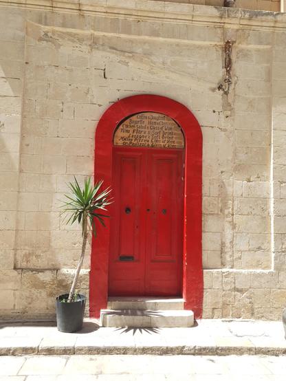 Eine rote Tür führt in ein Gebäude aus Sandstein. Sie ist geschlossen. Im oberen Türbogen steht eine Speisekarte auf Englisch geschrieben. Coffee & Doughnuts, Ham & Cheese, Pizza, Glass of  Wine ... Links neben der Tür steht eine Palme in einem schwarzen Blumentopf.
