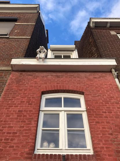 Photograph of a narrow red brick building facade looking up. On top of the eaves is a small unpainted ceramic sculpture of a man sitting on the edge of the roof with his legs dangling over, holding up and looking through binoculars straight on to the view behind the photographer. A dormer window is visible above the eaves. Two other taller brown brick buildings are on either side and the sky is bright blue with a few wispy clouds. Below the eaves in the centre of the frame is a window with white window frames and an arched top. Inside the window two ceramic human heads with painted hair are visible placed so they are looking out the window. Maastricht, Netherlands, 22 May 2024. Photo Cindy Kohtala