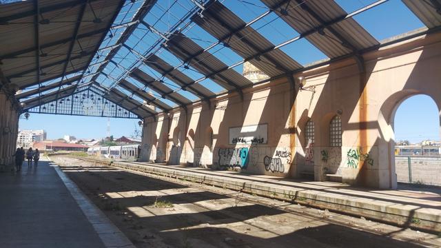 Ein altes, nicht mehr genutztes Bahnhofsgebäude. Im Dachgerüst scheinen Glasfenster zu fehlen, die beiden Gleise wurden entfernt. Es ist sonnig. Die Wände sind mit Graffiti bedeckt.