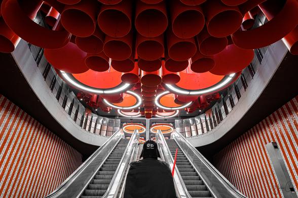 Self-portrait in a Darth Vador costume facing the futuristic background of the Pannenhuis metro station in Belgium, with a giant escalator ascending under a ceiling composed of red and orange giant tubes and lamps hanging.