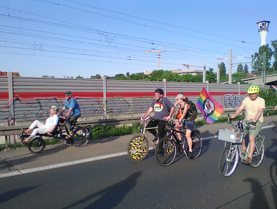 Radfahrende auf einer Schnellstraße, im Hintergrund der Qelleturm und die Bahnstrecke Nürnberg-Fürth mit Lärmschutzwand im ICE-Design, davor ein Tandem vorne halb liegender Mitfahrerin, zwei Radelnde mit XR-Zeichen am Rad bzw. einer Regenbogen-Antifa-Fahne