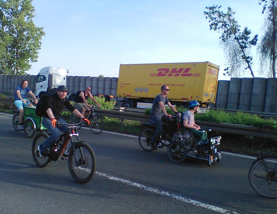 Radfahrer auf einer Schnellstraße, vorne ein Geländefahrrad mit dicken Reifen und Federgabel, ein Transport-Fahrrad mit Rollstuhlfahrer auf einer Ladefläche vor dem Lenker und ein Lasten-Dreirad, im Hintergrund ein gelber DHL-Laster auf der Gegenfahrbahn