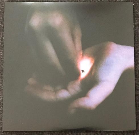 A close-up image of two hands, one hand is holding a small object while the other hand is positioned above it, creating a soft glow or light effect between them. The background is dark, emphasizing the hands and the light.