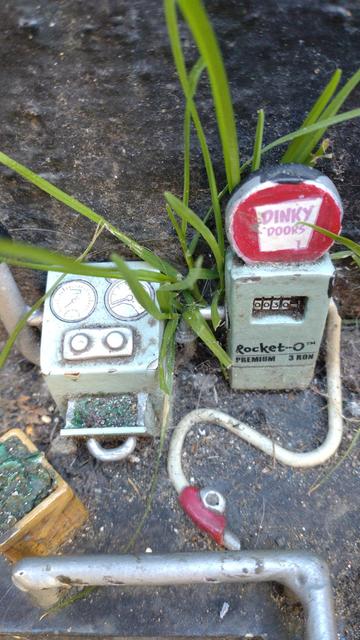 What appears to be a scaled down petrol station. There's a pump with a sign that says "Diny Doors" showing 30 units of fuel and "Rocket-O PREMIUM 3 RON" fuel, a box of raw materials and a machine for converting it into fuel. Several pipes run between the machines. The small scale is clear from the fact a plume of grass is sprouting from behind, overshadowing the tiny pump.
