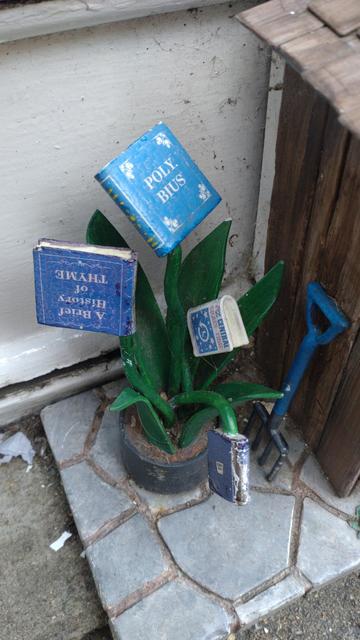 What appears to be a sculpted plant growing books. The books are titled "Poly. Bius", "A Brief History of THYME", "AZ CENTRAL SPACE-TIME" and one further book where the title can't be seen. A fork is stuck in the ground next to the plant pot, apparently leaning up against a shed with a crazy-paving floor. Everything is small in scale.