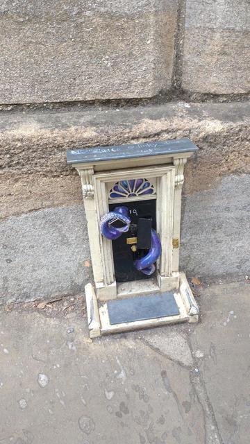 A tiny door, about 30 cms in height, leading in to the base of a much larger stone building. The door looks like the door to Number 10, Downing Street (the UK Prime Minister's official residence). Bits of the door have been forced from behind by a purple tentacled creature. Two of the tentacles have burst forth and are protruding from the holes where the panels have been pushed away.