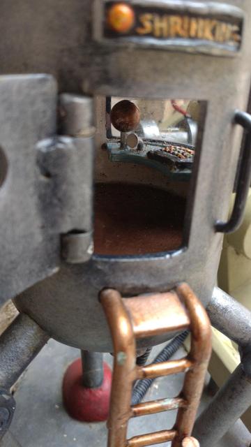 What appears to be a door in the side of a metal capsule on stilts. Stairs lead up to the open door. Above the door there's a light besides which is the word "SHRINKING". Inside the capsule a console can be seen with a couple of what appear to be levers. From the focus, this appears to be a small object.