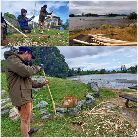 Two men crafting wooden poles by an ocean bay. Tools and wood shavings are scattered around the outdoor workspace.