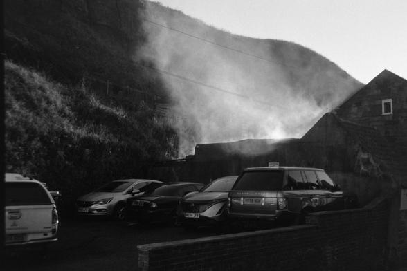 A black and white photograph showing smoke rising from a small structure. Some cars are parked in front.