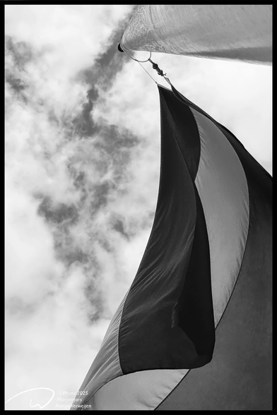 The Dutch flag seen from below hangs at half-mast on a flagpole against a partly cloudy sky.