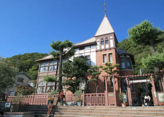 The image depicts a large, picturesque building with a distinctive architectural style. It features a combination of brick and wood elements, with a tower and steep roof. Surrounding the building are trees and a landscaped area, creating a warm and inviting atmosphere. The sky is clear and blue, adding to the overall aesthetic appeal of the scene. The building appears to be situated in a hilly area, and there are people visible, indicating that it's likely a public space or attraction.

Image Credits: Wikimedia / KishujiRapid / CC BY-SA 4.0