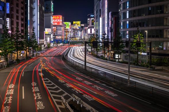 Long exposure shot on a buzzy main road of Shinjuku in Tokyo from a bridge over the road, at night. Only the light trails of the cars are visible on the road in the foreground due to the long exposure. Skyscrappers and buildings covered in colourful neons are recognisable in the background.