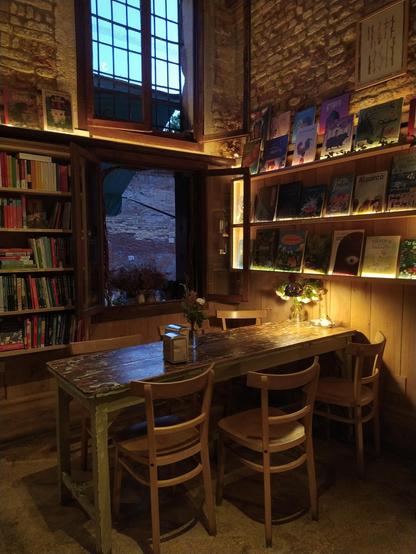 Foto von dem Haus von innen. Man sieht einen alten Tisch mit mehreren Holzstühlen, dahinter ist eine Wand mit zwei Fenstern übereinander. Die Decke ist so hoch, dass man sie nicht sieht. An sen Wänden sind Bücherregale. Alles liegt in einem orangenen Licht, am Ende des Tisches gibt eine Leselampe noch helleres gelbes Licht.