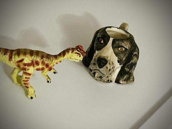 A green Dilophosaurus with brown spots looks at a little cup shaped like the head of a black and white English Springer Spaniel on a white backdrop.