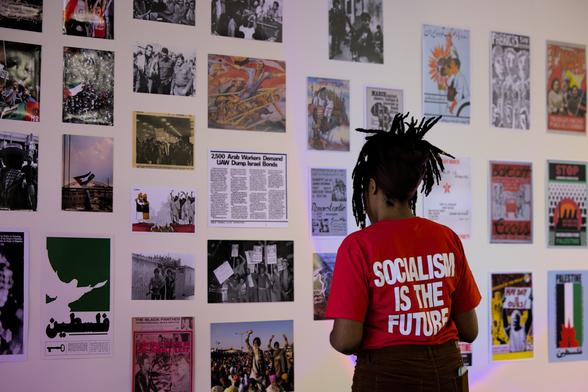A woman wearing a red shirt with the words "Socialism Is The Future" on the back, facing a wall covered with revolutionary art and photography.