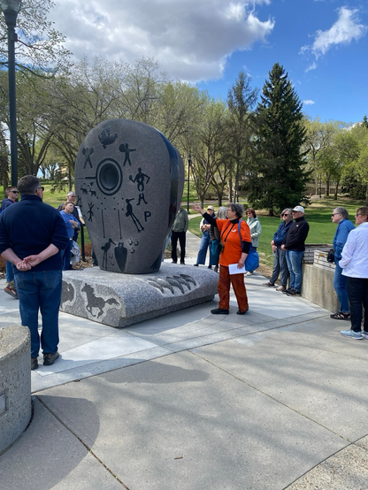 Senator Simons, in an orange Jane’s Walk t-shirt, points to the residential school memorial, in the Reconciliation Garden, while tour-takers look Senator .