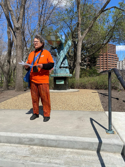 Sen Simons, in a Jane’s Walk orange t-shirt and batik orange pants stands in front of a statue near the front of the legislature, while wearing a headset mic and portable speaker.
