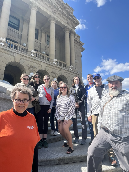 Paula Simons, left, stands with a group of her tour takers on the east steps of the Alberta legislature- with its sandstone pillars. To the far right of the photo, is Ian Hosler, a man with a grey beard and a flat tweed