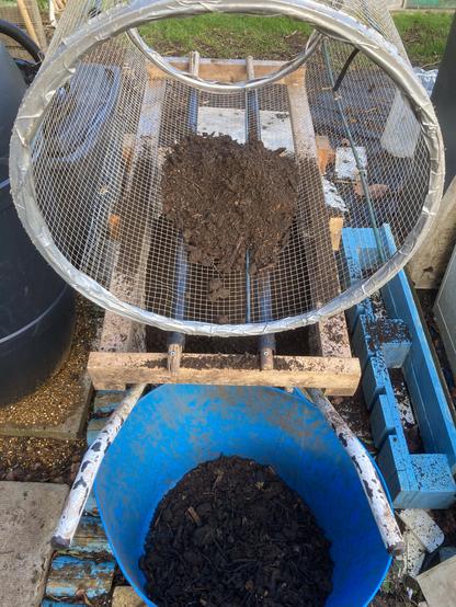 A compost trommel for quickly sifting compost. A wooden frame sits over a wheelbarrow. A wire mesh sheet is fitted to two old bicycle to make a tube shape. The mesh tube sits on the frame. The frame has two wooden poles with rollers to help spin the trommel tube. Compost is added inside the trommel tube which is turned by hand. The finer compost falls through the mesh into the wheelbarrow. The larger bits of compost fall into a trug between the wheelbarrow handles. These larger items can then go back into the compost heap or bin to further break down. The photo was taken last spring.