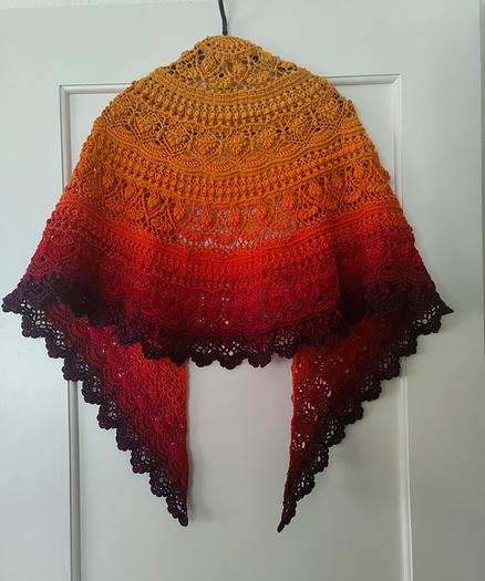 A crocheted shawl hanging on a white door. The shawl features intricate patterns and is gradient-colored, transitioning from a bright yellow at the top to a deep red and dark purple at the bottom. The edges of the shawl are scalloped, adding to its detailed design.
