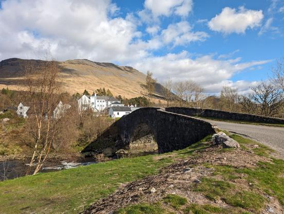 In the foreground, a green grassy area leading to a stone bridge which spans a small river. On the opposite side is a small village of white buildings with gray roofs. Behind the village is a large hill covered with brown grasses with darker patches. The sky is mostly cloudy with a bright blue patch in the top right corner.