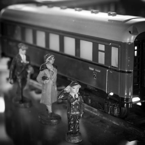 Von einer Modelleisenbahn sieht man schräg einen Waggon auf der rechten Seite. Vor dem Zug kann man drei kleine Personen sehen, die kleinste im Vordergrund salutiert.