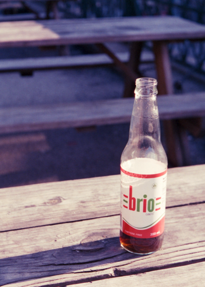 A clear glass bottle sits on a picnic table, a small amount of the caramel brown fluid remaining. The white label proclaims “brio” in green and red printing and graphics, reminiscent of the Italian flag.