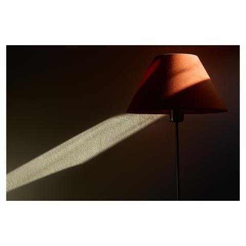 A table lamp with a rusty brown shade in an almost dark room. Sunlight from a window illuminates a small part of the wall behind the lamp, making it seem as if the light emanates from the lamp.