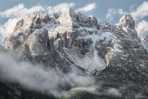An image of towering mountains from the North Italian Dolomites. The snow covered peaks surround a large cirque like a crown. Blue sky and some light greens from the forest below add color. And heavy clouds sweep into the valley from the sides of the image