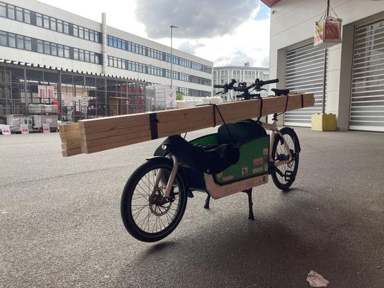 Foto zeigt Lastenrad mit jeweils 5 Dachlatten á 3 m Länge, jeweils rechts und links vom Lenk-/Sattelrohr, aufliegend auf der Kiste, 3 x verzurrt