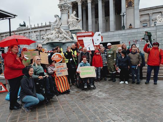 Ungefähr 25 Protestierende posieren vor dem Parlament auf dem Kopfsteinpflaster für ein Foto. Einige der Personen, manche mit und andere ohne Rollstuhl, halten Schilder hoch. Darauf stehen Slogans wie "Man ist nicht behindert, man wird behindert", "Ja zur Inklusion" oder "Genug geredet". Im Hintergrund sieht man abgesehen von dem Parlamentsgebäude auch eine nachgestellte Baustelle, die von den Organisationen zur Verdeutlichung der Anliegen aufgestellt wurde. Einige Personen tragen Bauhelme, um das Bild der künstlichen Baustelle zu vervollständigen.