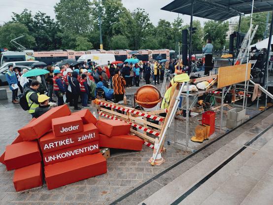 Das Foto zeigt den Blick von den Stufen des Parlaments auf die Straße, den Ring im ersten Bezirk in Wien. Im Blickfeld liegt die Kundgebung für Behindertenrechte. Die Protestierenden hören einer Person zu, die gerade auf der Bühne steht und eine Rede hält. Die Kundgebung wurde als Baustelle inszeniert. Es stehen Absperrungen und ein kleiner Zementmischer auf dem Platz vor dem Parlament. Außerdem haben die Organisator*innen riesige rote Blöcke in Form von Ziegelsteinen aufeinander gestapelt. Darauf zu lesen ist der Schriftzug: "UN Konvention. Jeder Artikel zählt!".