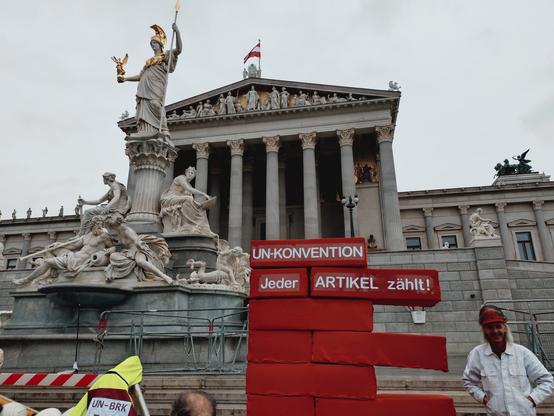 Es handelt sich um eine Nahaufnahme des Parlaments. Links im Bild ist der Brunnen mit seiner Statue vor dem Parlament zu sehen. Die Statue bildet die Pallas Athene, die Göttin der Weisheit, ab. Sie hält ein Zepter in in die Luft und ihr sitzen in Stein gemeißelte Personen zu Füßen, wie in Gedanken versunken. Vor der Statue und vor den Stufen zum Parlament haben die Demonstrierenden große rote Blöcke aufeinander gestapelt, die eine unvollständige Mauer bilden. Rechts neben den Blöcken steht eine Person. Sie lächelt in die Kamera und trägt eine weiße Jacke sowie einen roten Bauhelm. Der Stapel aus Klötzen ist ein paar Köpfe höher als sie. Auf den übergroßen Klötzen steht: "UN-Konvention. Jeder Artikel zählt."
