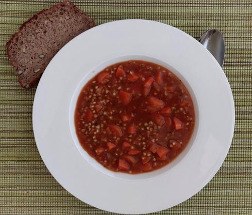Ein weißer Teller mit einer dunkelroten Suppe, in der Buchweizenkörner und Karottenstücke liegen. Daneben ein Kanten Brot und ein Löffel auf einem moosgrün-gestreiften Platzdeckchen.