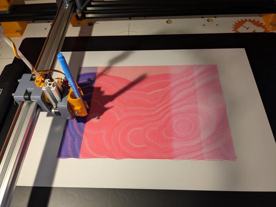 Top down view of a DIY pen plotter made from orange and grey 3d printed parts with shiny silver and black anodised aluminium rails. On the black plotting surface there is a piece of A3 white paper. The pen holder has a blue pen which is working across the sheet but already laid down is a previous pattern made up from small magenta/pink lines at different angles making swirly patterns